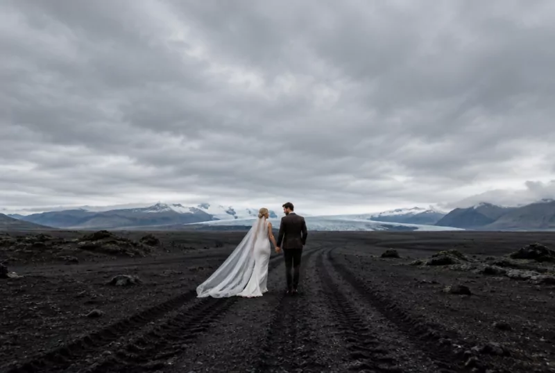 Brautpaar geht über eine schwarze Lavawüste in Island, am Horizont ein Gletscher unter tiefen Wolken