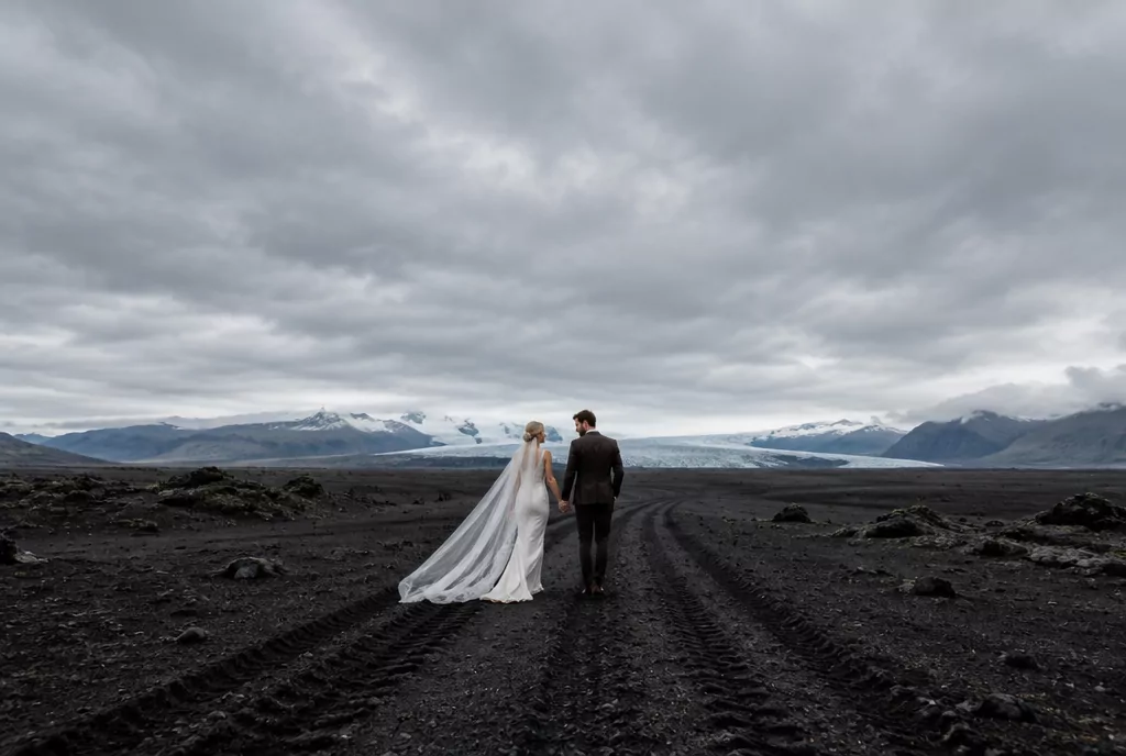 Brautpaar geht über eine schwarze Lavawüste in Island, am Horizont ein Gletscher unter tiefen Wolken